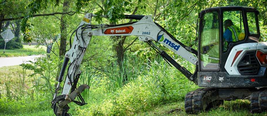Man in high-vis vest driving an MSD backhoe through tall grasses and trees